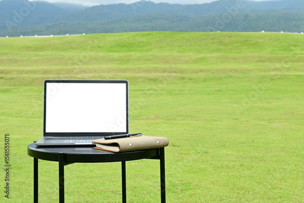 Obraz A laptop is open on a table in a grassy field. The grass is green and the sky is cloudy. The scene is peaceful and serene, with the laptop