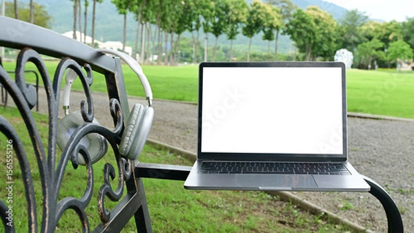 Obraz A laptop is sitting on a bench in a park. The laptop is open and the screen is blank. The bench is located in a grassy area with trees in the background.