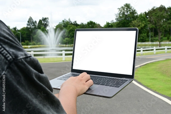 Obraz A person is holding a laptop in a park. The laptop screen is blank, and the person is typing on it.