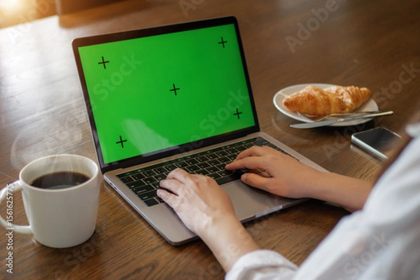 Fototapeta laptop blank green screen mockup. woman using laptop computer, hand typing on keyboard at home for work with hot coffee, croissant bakery and mobile phone on the wooden table in the morning.