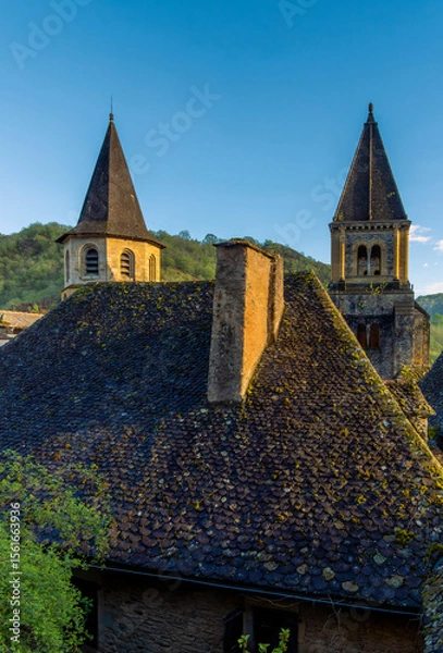 Fototapeta Twin spires and ancient tiled roofs and historic towers of the Abbey of Sainte-Foy, in the medieval village of Conques, France, illuminated by the golden light of sunset.
