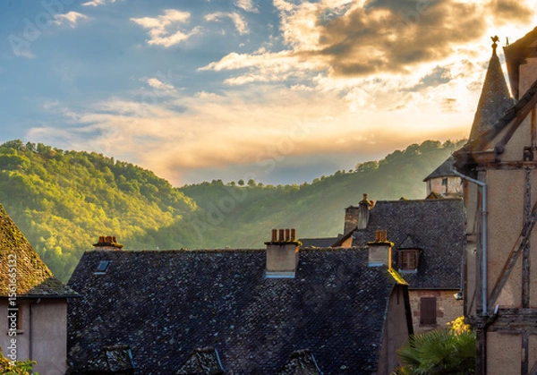 Fototapeta Golden hour over the rooftops of the French village of Conques, with rustic medieval houses and mountains illuminated by the sunset light under a spectacular sky with backlit clouds.
