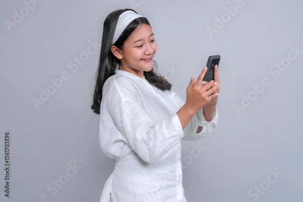 Fototapeta Cheerful young Asian woman in a white shirt and headband smiling while using a smartphone, looking at the screen with joy and excitement, isolated on a plain gray background.