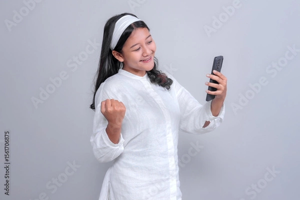 Fototapeta Asian woman wearing a white shirt and headband, holding smartphone while smiling and raising her fist in a happy, victorious pose, isolated on grey background, studio shot.