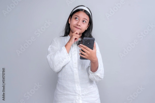 Fototapeta Thoughtful young Asian woman in a white shirt and headband holding a smartphone while touching her chin, appearing curious or uncertain, isolated on a plain studio background.