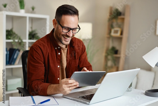 Obraz Happy entrepreneur working from home using laptop and tablet smiling
