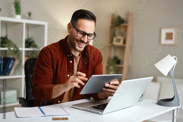 Obraz Smiling freelancer working from home using laptop and tablet