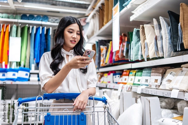 Obraz A young asian woman pushes a shopping cart and gazes at her mobile phone, browsing through a variety of bedding items like pillows and sheets in a vibrant department store.