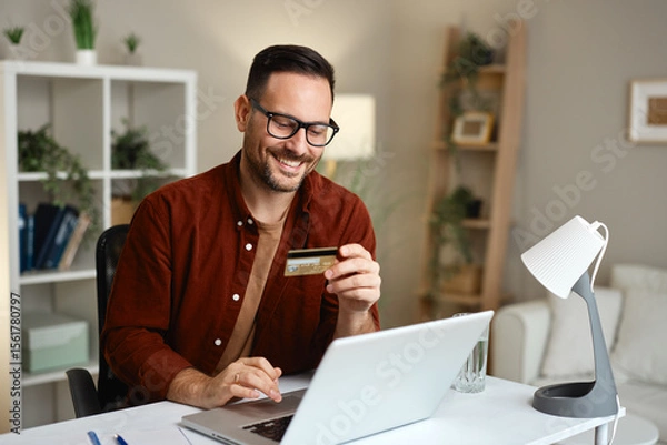 Obraz Smiling man using laptop and credit card for online shopping at home office