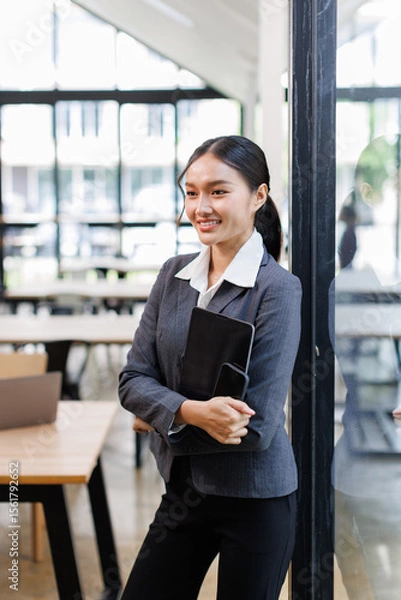 Obraz Beautiful asian business woman in a suit standing and holding tablet confidently with arms folded, isolated on white background,
