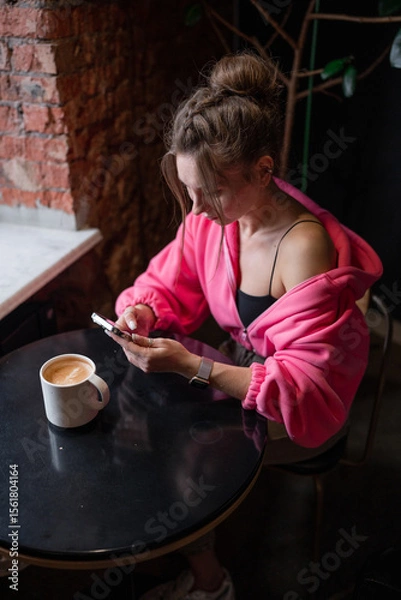 Fototapeta A stylish woman enjoying her coffee while scrolling on her phone in a warm cafe setting.