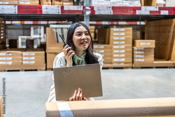 Obraz lively warehouse, dedicated young asian woman meticulously checks inventory details on her laptop while engaged in a conversation for radio communication, ensuring smooth operations and accuracy