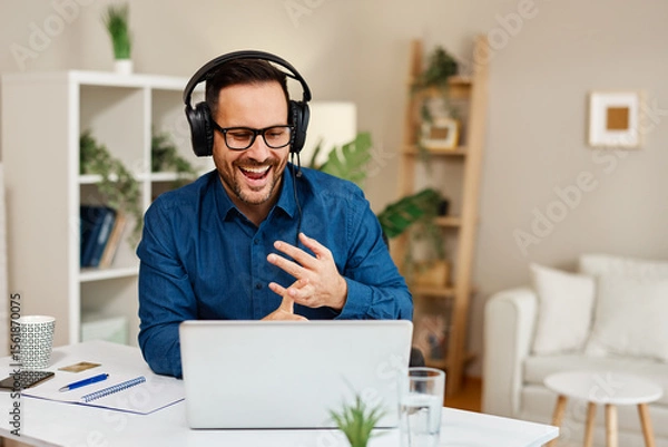 Obraz Happy businessman wearing headphones having a video call on laptop and gesturing at home