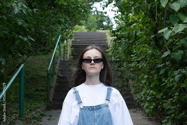Fototapeta A young individual wearing a white shirt and denim overalls confidently ascends steps in a lush green area. Sunlight filters through the trees, creating a vibrant atmosphere