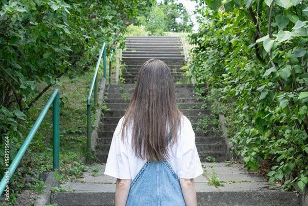 Fototapeta A young woman ascends stone steps bordered by lush greenery in a serene park. Sunlight filters through the leaves, creating a peaceful atmosphere