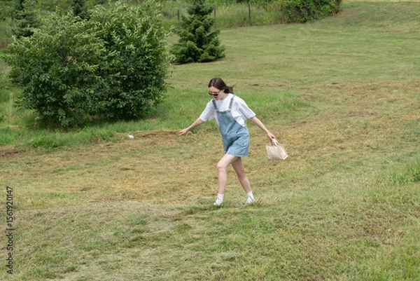 Fototapeta A young woman wearing denim overalls and sunglasses enjoys a warm, sunny day as she walks through a lush, grassy field. She carries a small bag while exploring the outdoors
