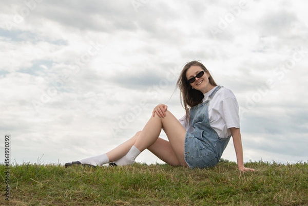 Fototapeta A young woman sits casually on a grassy hill wearing denim overalls and sunglasses. She smiles while enjoying the fresh air under a partly cloudy sky during a leisurely afternoon