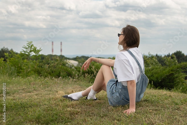 Fototapeta A young woman sits comfortably on a grassy hill, enjoying a moment of relaxation while gazing at the horizon. The cloudy sky adds a serene touch to the industrial view