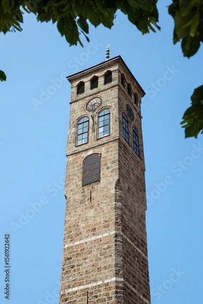 Fototapeta Historic Clock Tower Against a Clear Blue Sky Framed by Green Leaves.Sarajevo Uhrturm(Clock Tower)