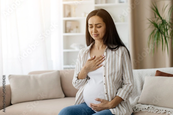 Fototapeta Pregnant woman feeling discomfort, touching chest with one hand and belly with the other, expecting female with concerned facial expression, sitting on couch in home interior, copy space