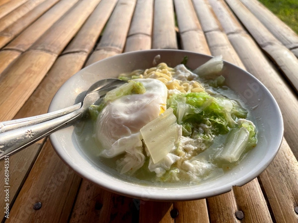Fototapeta Hand movements holding spoons and forks, stirring a bowl of delicious steaming instant noodles, complemented by fried eggs and chopped green vegetables such as cabbage. The bowl is placed on a bamboo 