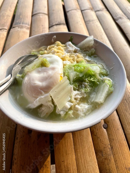 Fototapeta Hand movements holding spoons and forks, stirring a bowl of delicious steaming instant noodles, complemented by fried eggs and chopped green vegetables such as cabbage. The bowl is placed on a bamboo 
