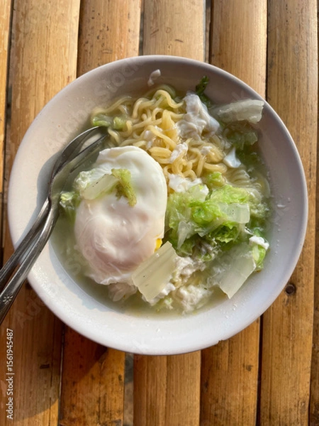 Fototapeta Hand movements holding spoons and forks, stirring a bowl of delicious steaming instant noodles, complemented by fried eggs and chopped green vegetables such as cabbage. The bowl is placed on a bamboo 