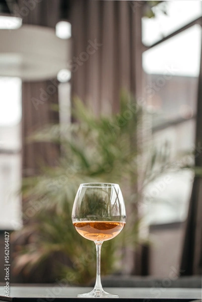 Obraz A single glass of amber wine stands elegantly on a dark table, with a softly blurred background of indoor greenery and warm light