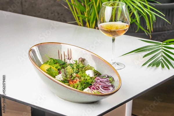 Obraz A fresh Greek-style salad with feta, red onion, and greens served in a ceramic bowl next to a glass of white wine on a modern white table