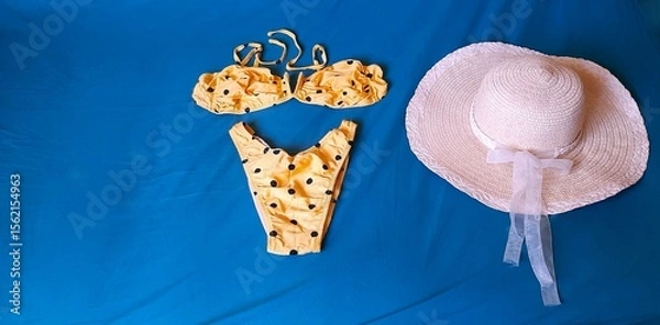 Fototapeta Yellow bikini set with black polka dots and a straw hat with white ribbon, laid out on a vibrant blue background. Summer preparations, with copy space