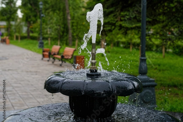 Obraz A close-up view of a water jet emerging from a metal fountain in a summer city park, with benches and a paved alley in soft focus, evoking cool relaxation. Photo. High resolution