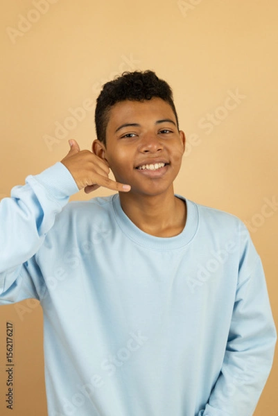 Fototapeta Vertical image of a cheerful brown-skinned Latino teenager wearing a pastel blue sweatshirt making a call gesture with his hand on a light brown background with copy space