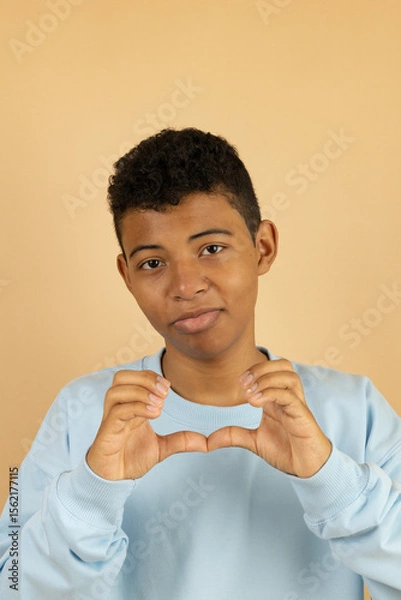 Fototapeta Vertical image of a dark skinned Latino teenager wearing a pastel blue sweatshirt making a broken heart with his hands on a light brown background with copy space, young man making a broken heart