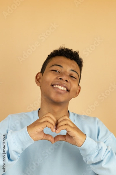 Fototapeta Vertical image of a cheerful brown skinned teenager wearing a pastel blue sweatshirt making a heart gesture with his hand on a light brown background with copy space, feeling of love with his hands