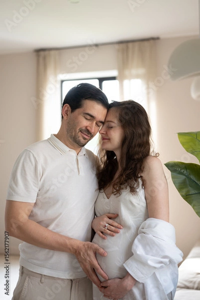 Obraz Happy man and pregnant woman gently touching belly with hands, enjoying pregnancy time together in cozy bedroom
