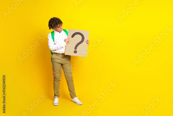 Fototapeta Portrait of a schoolboy holding a question mark sign against a yellow background to symbolize curiosity and learning