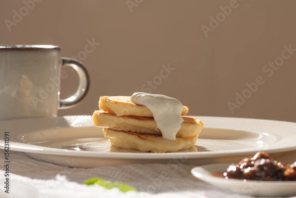 Obraz Pancakes with cream and jam on white plate with cup of coffee during breakfast, enjoying morning sunlight
