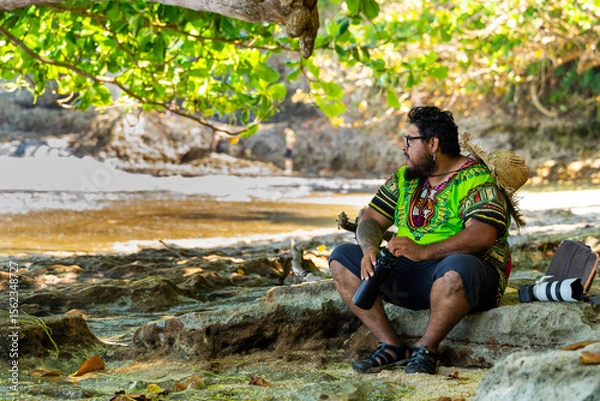 Fototapeta Local Costa Rican man in a Caribbean shirt resting under a tree with a recyclable water bottle on a beach in Cahuita National Park, Costa Rica
