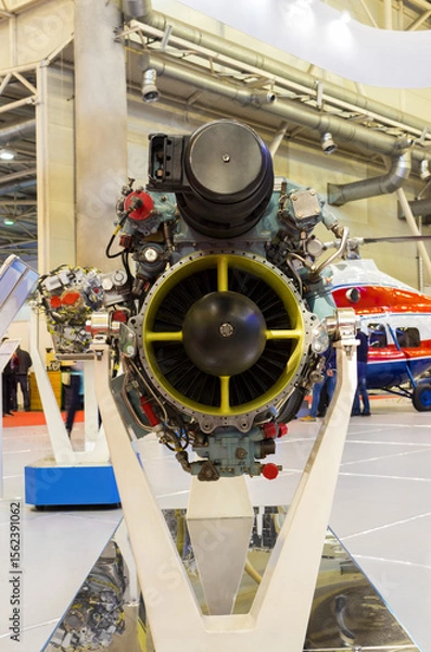 Fototapeta Helicopter gas turbine engine on display at aviation exhibition. Engine is mounted on stand, showcasing its intricate mechanical components and turbine blades. Colorful helicopter in background