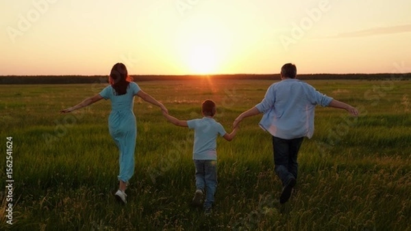 Obraz Father mother child son running together holding hands on green meadow at sunset. Family, parents child playing together in sunny park. Slow motion. Picnic dad mom child active recreation in nature