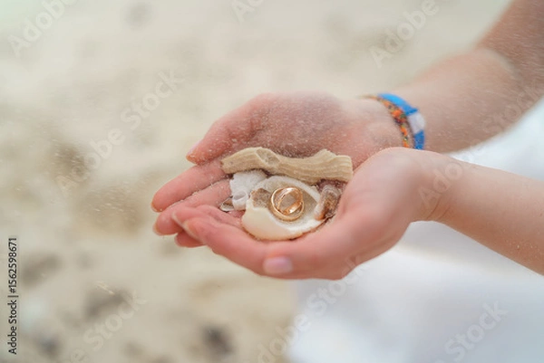 Fototapeta Wedding rings and small shells in the hands of a girl.