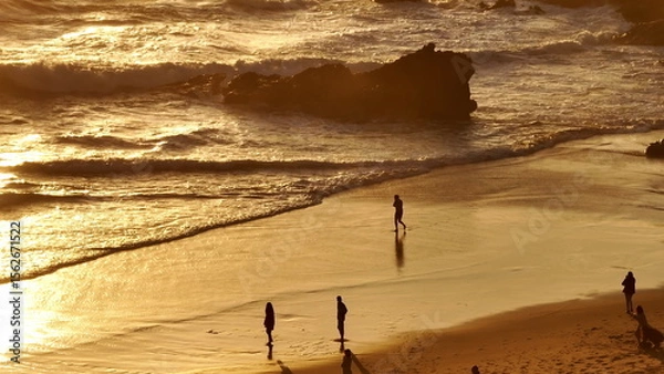 Obraz Lisbon, Portugal: Golden sunlight reflecting on wet orange sand, tourists people walk along the sunset beach, creating a warm and inviting relaxing joy atmosphere. Summer travel vacation background