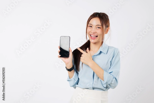 Fototapeta Smiling Asian woman holding a mobile phone, pointing a finger to its blank display. She is happy and excited, showing her new phone. isolated studio white background