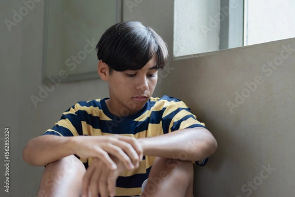 Fototapeta A teenage boy sits alone indoors near glass window with a thoughtful, melancholic expression, evoking feelings of solitude and introspection.