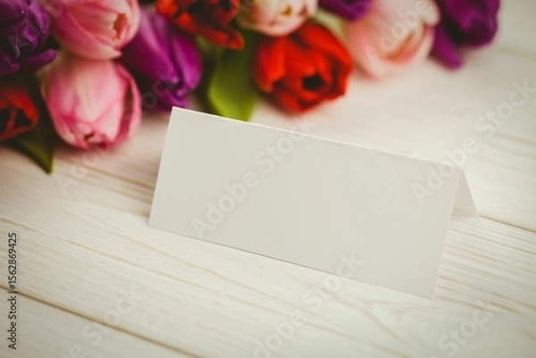 Obraz folded white place card is sitting on whitewashed tabletop, showing colorful tulip bouquet backdrop