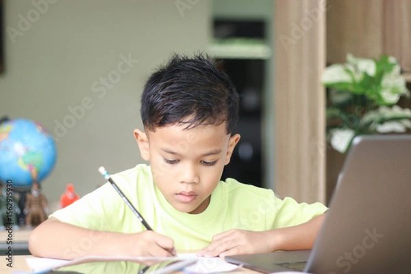 Fototapeta Young boy studying and doing homework at home with notebook and laptop. Concept of distance learning, education, homeschooling, and online classes for children.