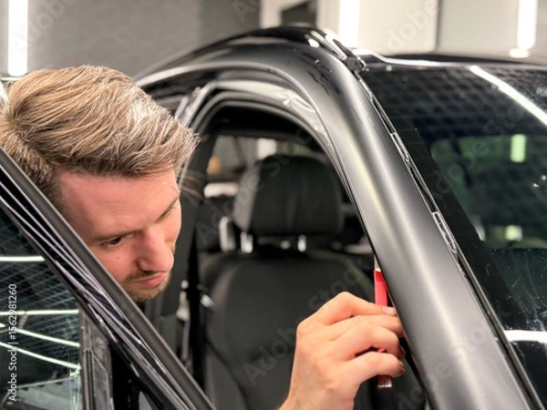 Obraz A car wrapping specialist applies a polyurethane film to the car. Detailer smoothes PPF. Car wrapping, detailing, tuning and styling. Selective focus. Car detailing and wrapping workshop. 
