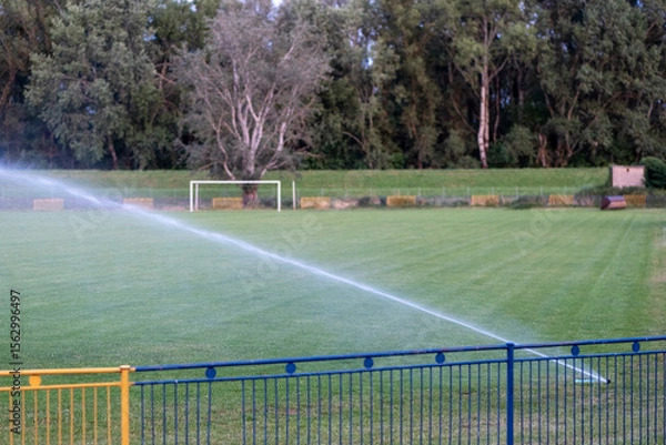 Fototapeta Sprinkler irrigating green sports field to prevent drought damage, showing increasing need for artificial watering due to climate change and dry conditions.