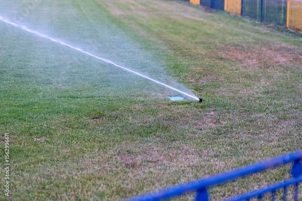 Fototapeta Automatic sprinkler irrigating sports field with dry grass patches, highlighting the need for watering to prevent drought damage and showing impact of climate change on green spaces.