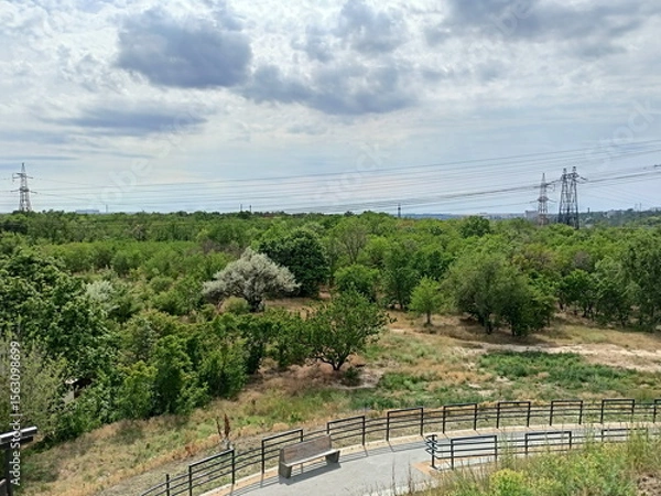 Fototapeta Panorama from the top of Unity Hill to the young green forest of Khortytsia Island against the background of a cloudy sky illuminated by the rays of the midday sun.